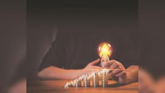 A hand holds a glowing lightbulb above a series of stacked coins arranged in a progressive, ascending fashion, symbolizing growth and investment potential.