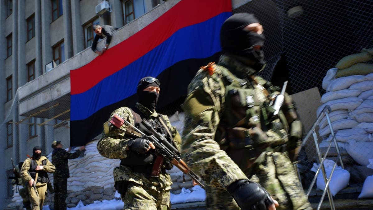 Armed individuals wearing masks and military attire walk past sandbags and a large flag featuring black, red, and blue stripes, as one person adjusts the flag above them.