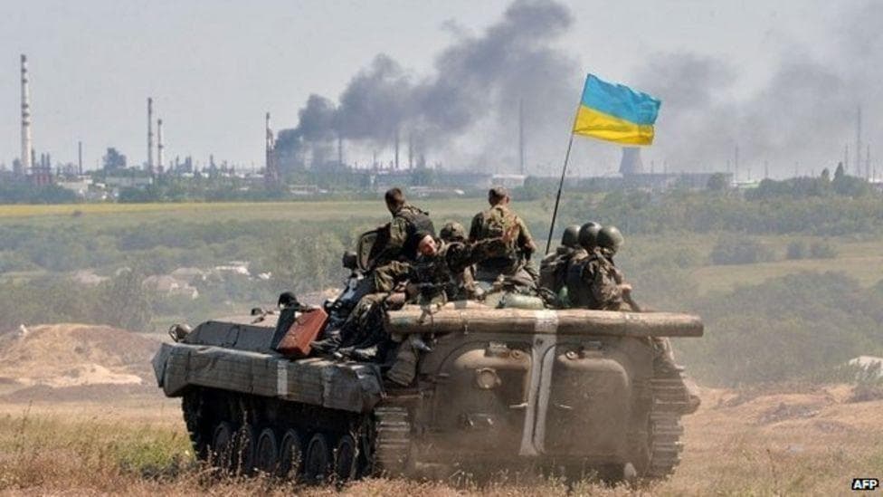 Ukrainian soldiers are stationed on a tank displaying the national flag, with industrial structures and smoke in the background, indicative of a conflict zone.