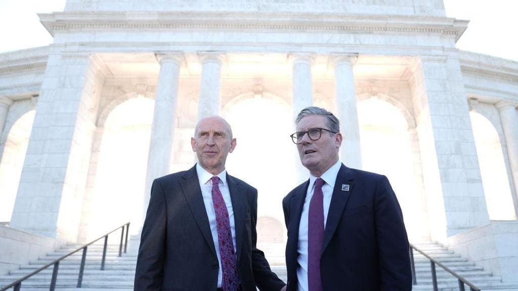 John Healey and another official stand on steps in front of a large monument, engaged in conversation.