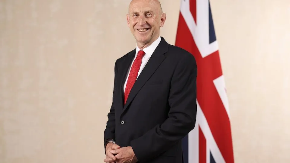 John Healey, wearing a black suit with a red tie, stands smiling beside a Union Jack flag.
