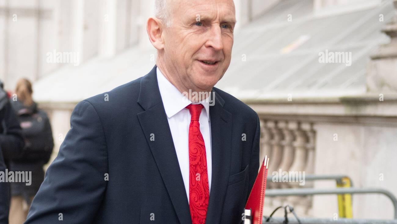 John Healey is smiling while wearing a dark suit and a red tie, holding red folders, as he walks past a building.