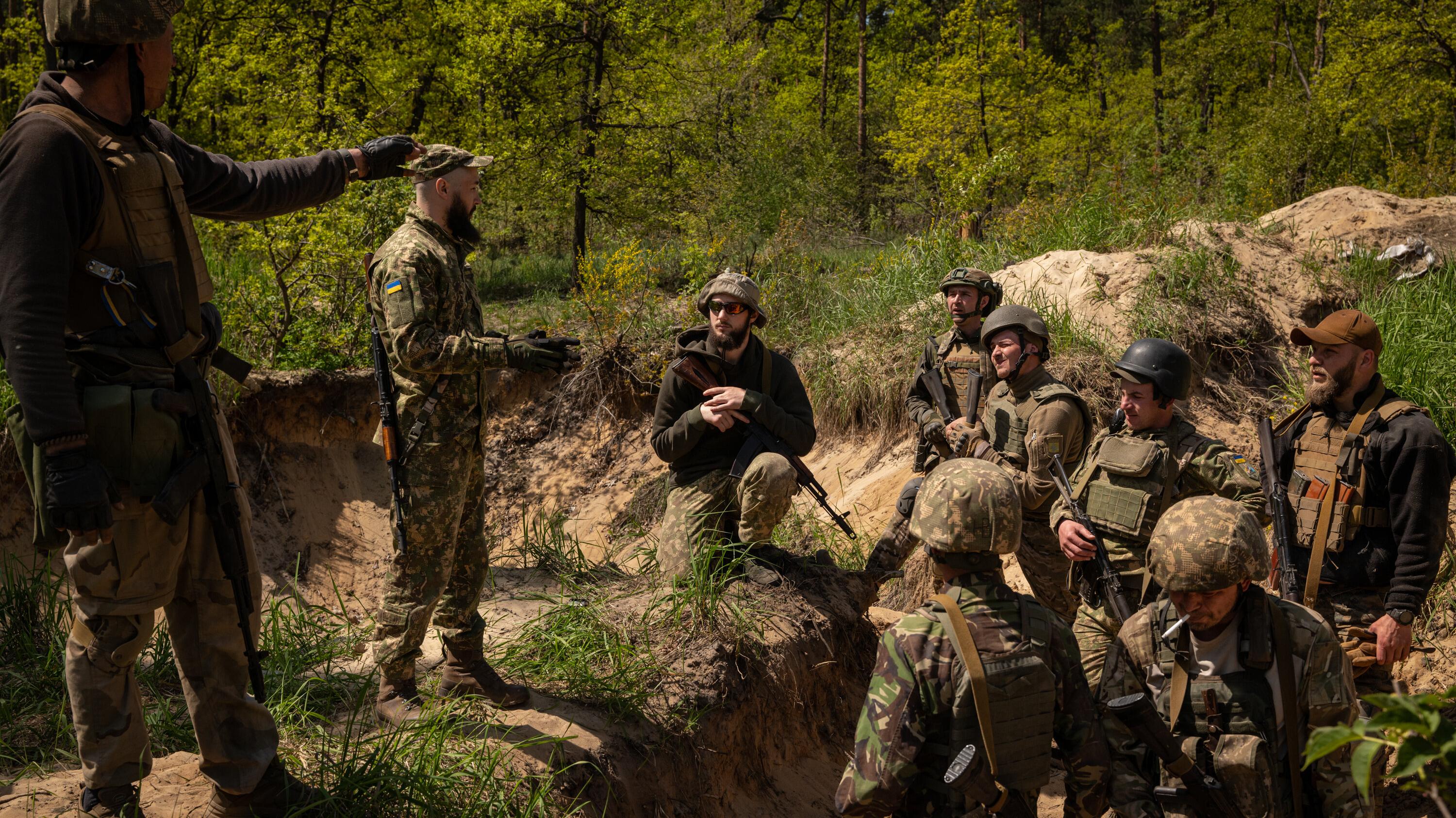 Ukrainian soldiers are engaged in a discussion amidst a wooded area, with some individuals standing and others seated in a trench, all equipped with weapons and military gear.