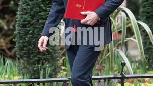 John Healey, the UK Defence Secretary, is walking confidently while holding a red folder, dressed in a formal suit with a tie, against a backdrop of carefully maintained greenery.